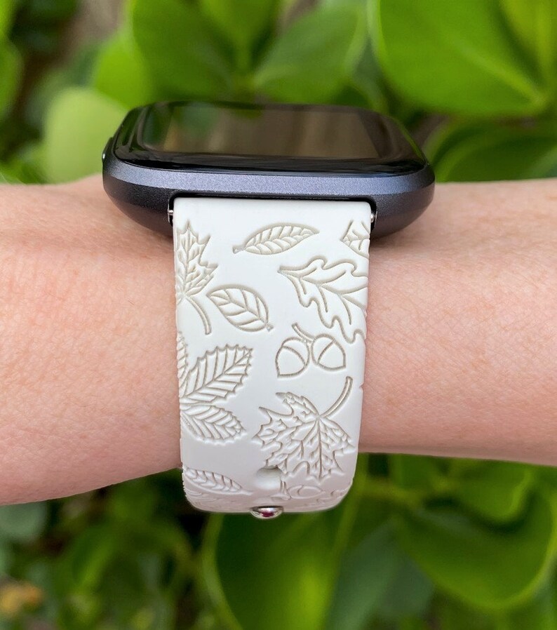 May include: A smartwatch with a white band featuring a leaf and acorn pattern. The band is made of a flexible material and is attached to a dark gray watch face. The design evokes an autumn theme.