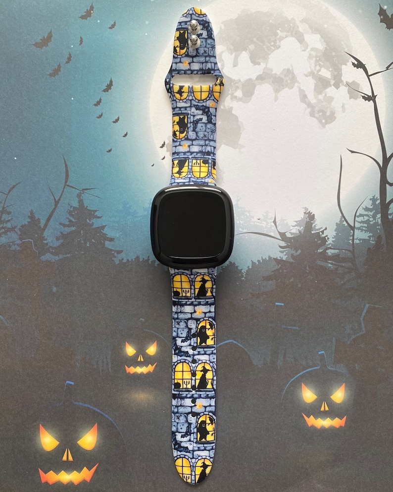 May include: A black smartwatch with a blue and yellow Halloween-themed band. The band features a repeating pattern of brick walls, windows, and silhouettes of witches and cats.