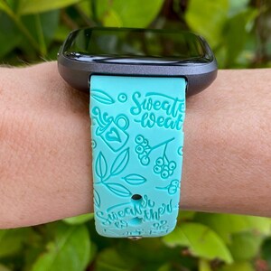 Pu&ograve; includere: Un cinturino per smartwatch turchese con un quadrante nero. Il cinturino ha un design bianco con il testo "Sweater Weather" e altre piccole illustrazioni.