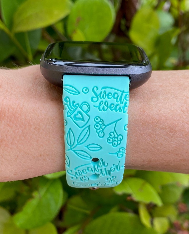 Pu&ograve; includere: Un cinturino per smartwatch turchese con un quadrante nero. Il cinturino ha un design bianco con il testo "Sweater Weather" e altre piccole illustrazioni.