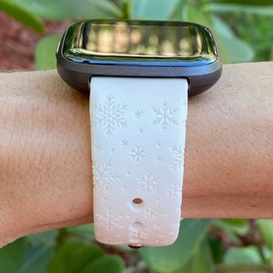 K&ouml;nnte beinhalten: Wei&szlig;es Silikon-Armband mit Schneeflockenmuster. Das Armband ist an einer schwarzen Smartwatch befestigt.