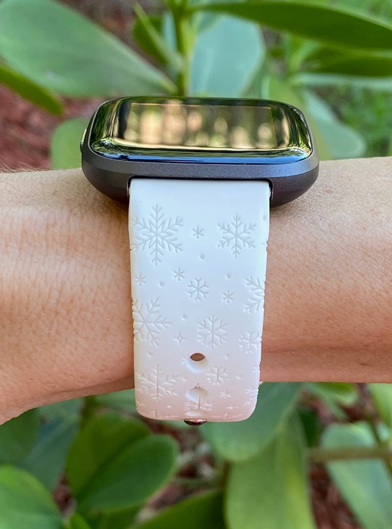 K&ouml;nnte beinhalten: Wei&szlig;es Silikon-Armband mit Schneeflockenmuster. Das Armband ist an einer schwarzen Smartwatch befestigt.