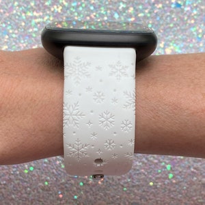 Pu&ograve; includere: Cinturino bianco per smartwatch con motivo a fiocco di neve. Il cinturino &egrave; in silicone e ha una fibbia in metallo nero.