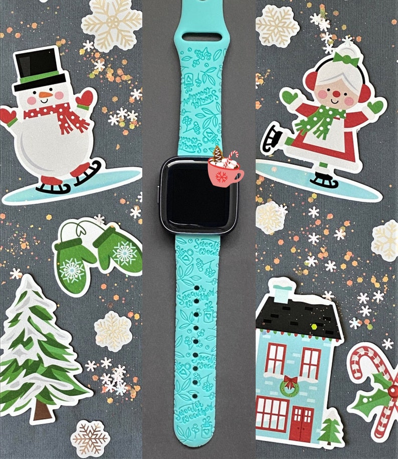 Pu&ograve; includere: Un cinturino per smartwatch blu turchese con un fiocco di neve bianco e un design "Sweater Weather". Il cinturino &egrave; su uno sfondo grigio con vari adesivi a tema invernale, tra cui un pupazzo di neve, una casa e una canna di zucchero.