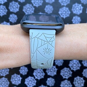 May include: A gray smartwatch with a spiderweb and spider design on the band.