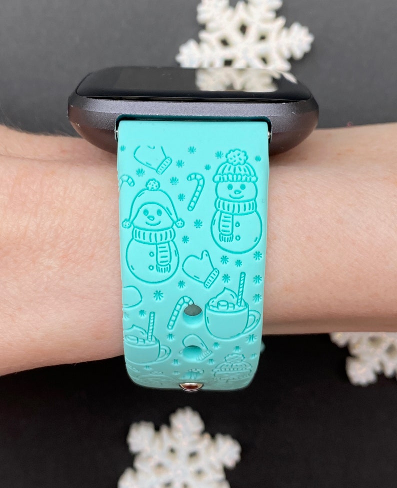 May include: A turquoise blue smartwatch band with a winter-themed design featuring snowmen, candy canes, and hot chocolate.