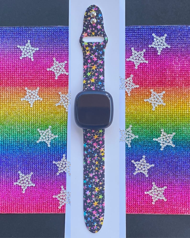 May include: A black smartwatch band with a colorful star pattern. The band is made of silicone and has a black buckle. The band is shown against a rainbow background.
