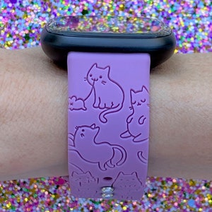 May include: A purple silicone watch band with a design of multiple cats. The band has two clear rhinestones on the bottom.