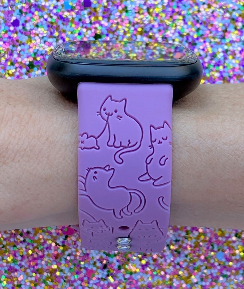 May include: A purple silicone watch band with a design of multiple cats. The band has two clear rhinestones on the bottom.