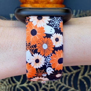 Peut inclure: Une montre intelligente noire avec un bracelet sur le th&egrave;me d'Halloween. Le bracelet pr&eacute;sente des marguerites orange et blanches, des fant&ocirc;mes blancs et des toiles d'araign&eacute;es sur un fond noir. La montre est attach&eacute;e &agrave; un poignet.