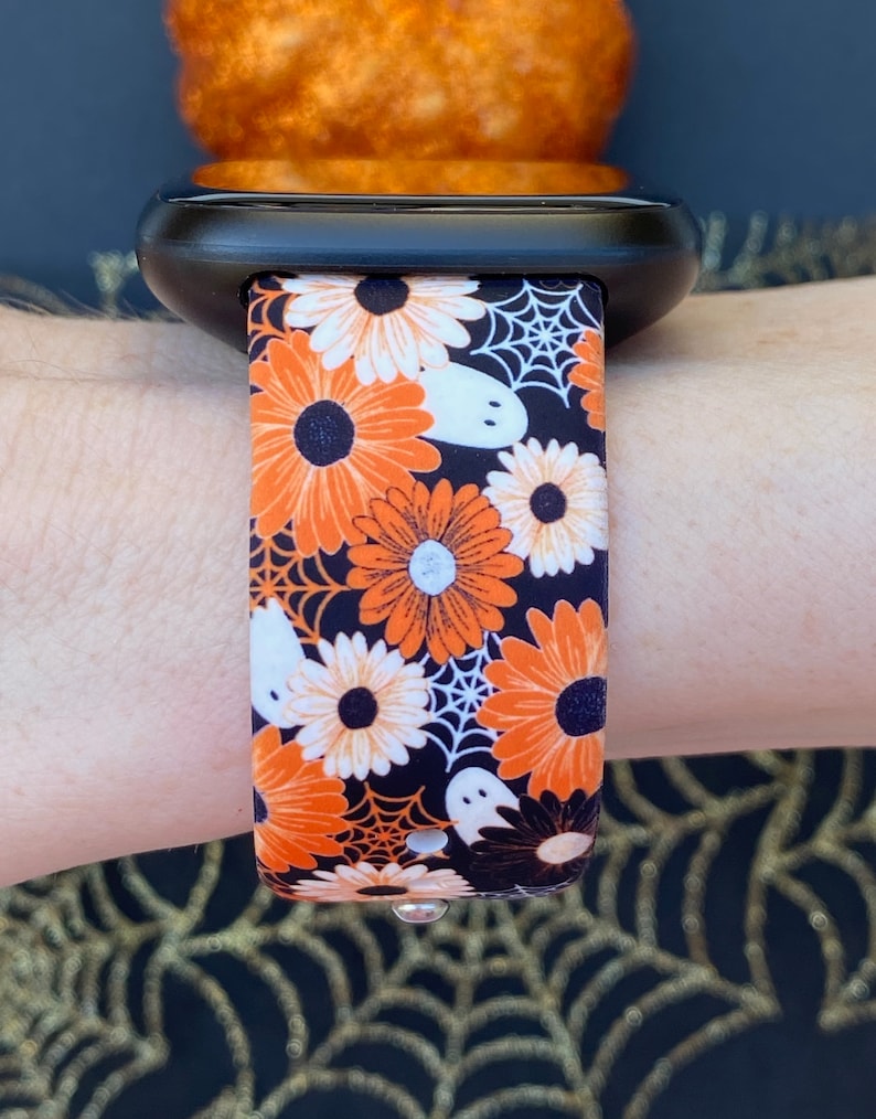 Peut inclure: Une montre intelligente noire avec un bracelet sur le th&egrave;me d'Halloween. Le bracelet pr&eacute;sente des marguerites orange et blanches, des fant&ocirc;mes blancs et des toiles d'araign&eacute;es sur un fond noir. La montre est attach&eacute;e &agrave; un poignet.