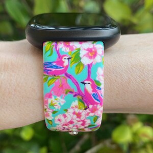 K&ouml;nnte beinhalten: Eine schwarze Smartwatch mit einem t&uuml;rkisfarbenen, rosa und wei&szlig;en Armband mit floralem Muster und rosa V&ouml;geln. Das Armband hat eine silberne Schlie&szlig;e.