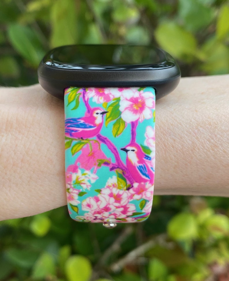 K&ouml;nnte beinhalten: Eine schwarze Smartwatch mit einem t&uuml;rkisfarbenen, rosa und wei&szlig;en Armband mit floralem Muster und rosa V&ouml;geln. Das Armband hat eine silberne Schlie&szlig;e.