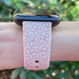 May include: A pink silicone watch band with a floral pattern. The band is designed for a smartwatch and has a small hole on the side.