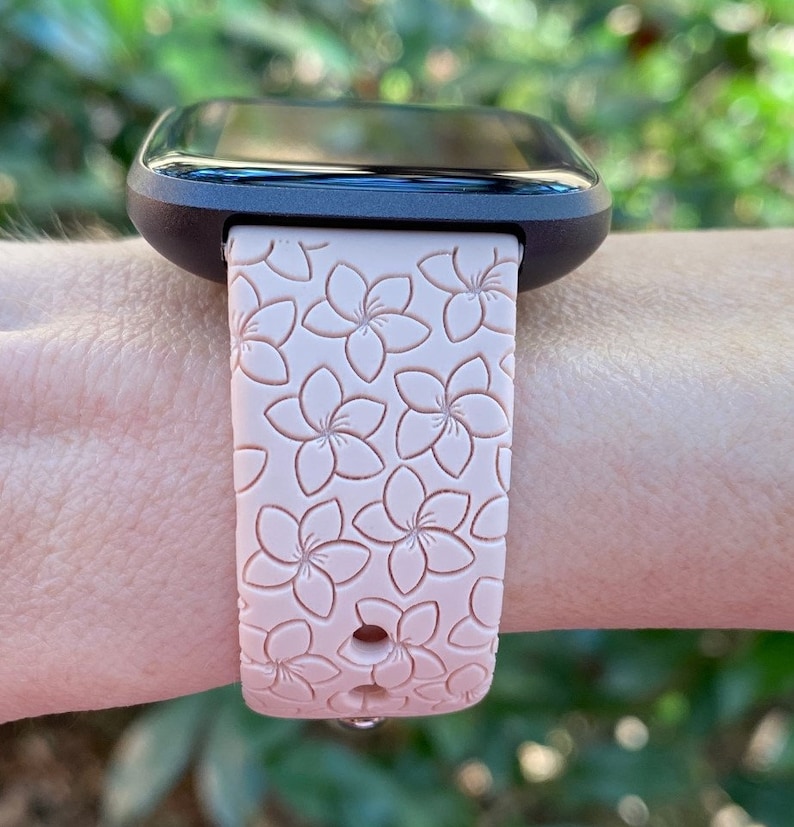 May include: A pink silicone watch band with a floral pattern. The band is designed for a smartwatch and has a small hole on the side.