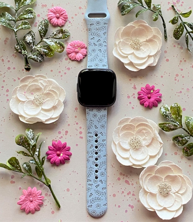 K&ouml;nnte beinhalten: Ein hellblaues Armband mit einem wei&szlig;en Blumenmuster. Das Armband ist an einer Smartwatch mit schwarzem Zifferblatt.