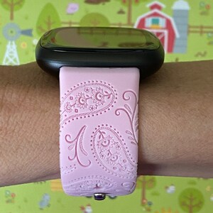 May include: A pink silicone watch band with a paisley pattern. The band is designed for a smartwatch and has a black watch face.