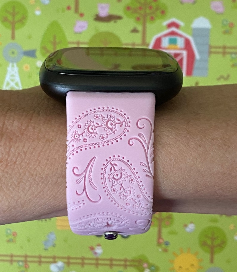 May include: A pink silicone watch band with a paisley pattern. The band is designed for a smartwatch and has a black watch face.