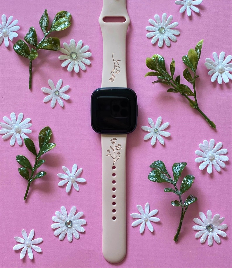 May include: A light pink smartwatch band with a floral design. The band is adorned with delicate brown floral illustrations. The watch face is black. The background is pink, with white daisy-shaped decorations and green leafy accents.