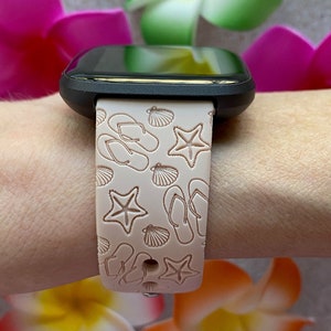 May include: A black smartwatch with a pink silicone band featuring a white embossed design of seashells, flip-flops, and starfish.
