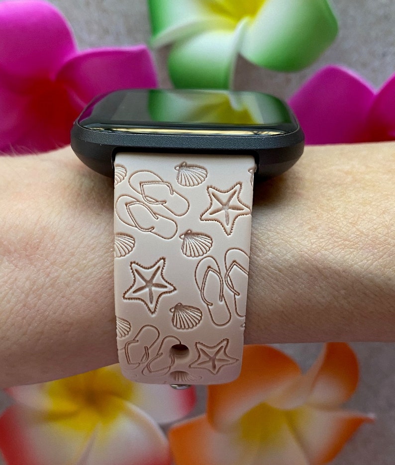 May include: A black smartwatch with a pink silicone band featuring a white embossed design of seashells, flip-flops, and starfish.
