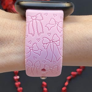 May include: A light pink watch band with a bow and heart pattern. The band is made of a soft material and is designed to fit a smartwatch. The band is shown on a wrist.