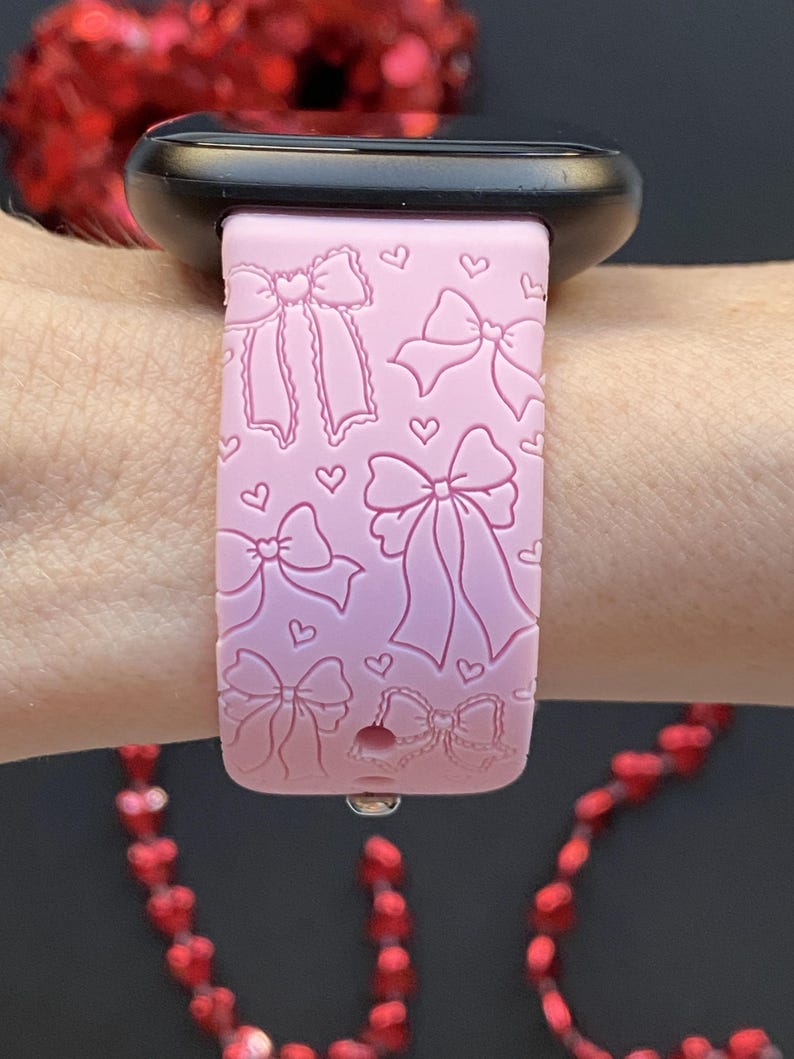 May include: A light pink watch band with a bow and heart pattern. The band is made of a soft material and is designed to fit a smartwatch. The band is shown on a wrist.