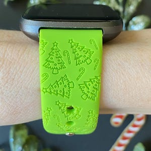May include: A bright green watch strap featuring a Christmas tree and candy cane design. The strap is attached to a silver and black smartwatch. The pattern is embossed on the strap.
