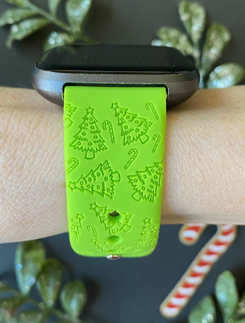 May include: A bright green watch strap featuring a Christmas tree and candy cane design. The strap is attached to a silver and black smartwatch. The pattern is embossed on the strap.