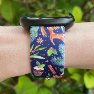 May include: A black smartwatch with a festive Christmas-themed band. The band features a blue background with green pine branches, red berries, cinnamon sticks, and a deer. A cupcake with red frosting and a holly sprig is also visible.