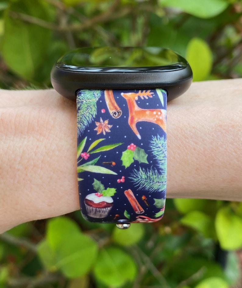 May include: A black smartwatch with a festive Christmas-themed band. The band features a blue background with green pine branches, red berries, cinnamon sticks, and a deer. A cupcake with red frosting and a holly sprig is also visible.