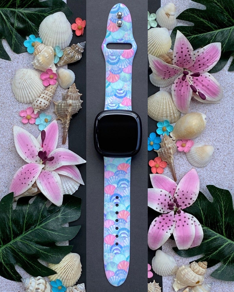 K&ouml;nnte beinhalten: Ein hellblaues, rosa und wei&szlig;es Armband mit Muschel-Muster. Das Armband ist aus Silikon und f&uuml;hlt sich angenehm weich an. Es ist kompatibel mit der Apple Watch Series 4 und neuer.