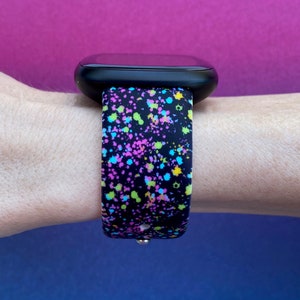May include: A black smartwatch band with a colorful splatter paint design. The band is made of silicone and has a black background with pink, blue, green, and yellow paint splatters.