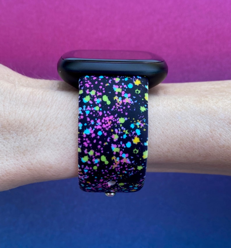 May include: A black smartwatch band with a colorful splatter paint design. The band is made of silicone and has a black background with pink, blue, green, and yellow paint splatters.
