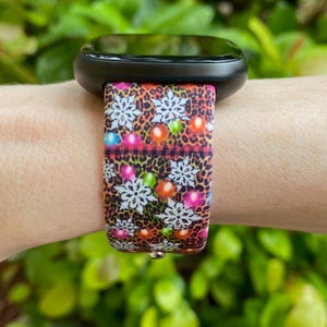 May include: A black smartwatch with a colourful Christmas lights and snowflake patterned band. The band is made of silicone and has a leopard print background.