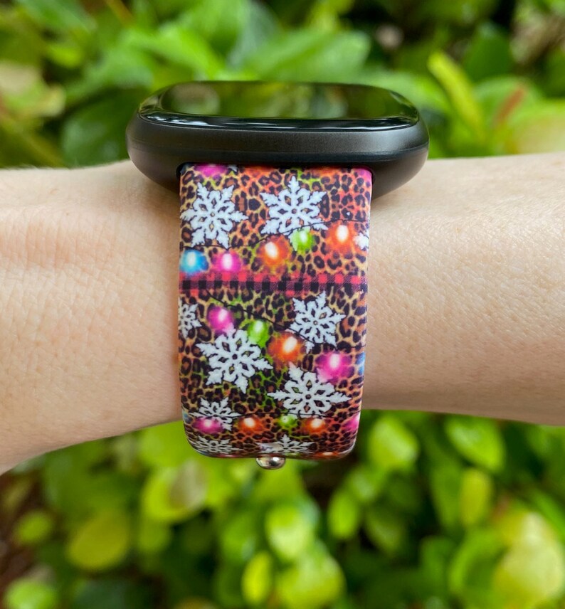 May include: A black smartwatch with a colourful Christmas lights and snowflake patterned band. The band is made of silicone and has a leopard print background.