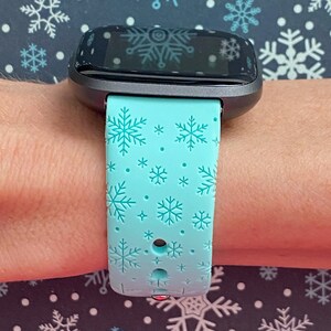 May include: A teal blue fitness tracker band with a snowflake pattern. The band is made of silicone and has a black buckle.