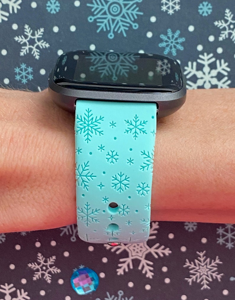 May include: A teal blue fitness tracker band with a snowflake pattern. The band is made of silicone and has a black buckle.