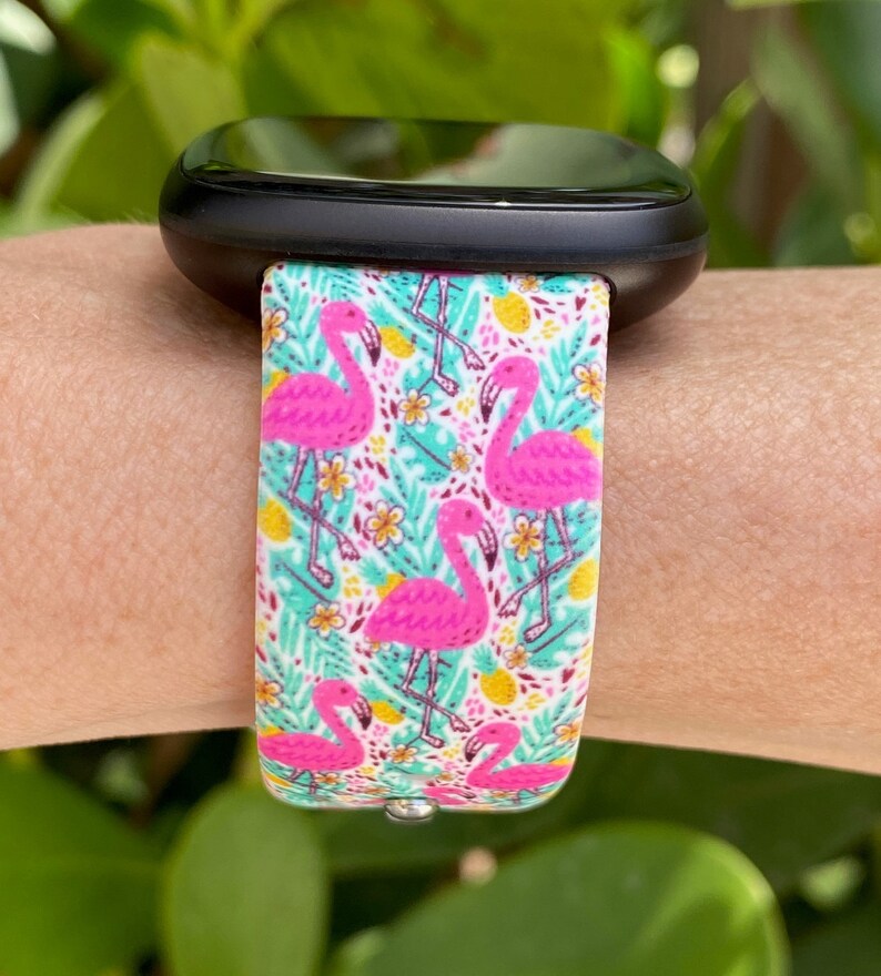 May include: A black smartwatch with a white and pink flamingo print band. The band features pink flamingos, green leaves, and yellow pineapples on a white background.