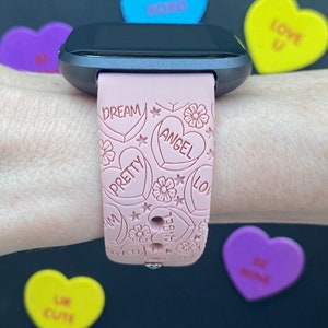 May include: A pink smartwatch strap with a heart design, featuring the words "Dream," "Angel," and "Pretty." The strap has floral and star details. Around the watch are colourful heart-shaped sweets with phrases such as "XOXO" and "Be Mine."