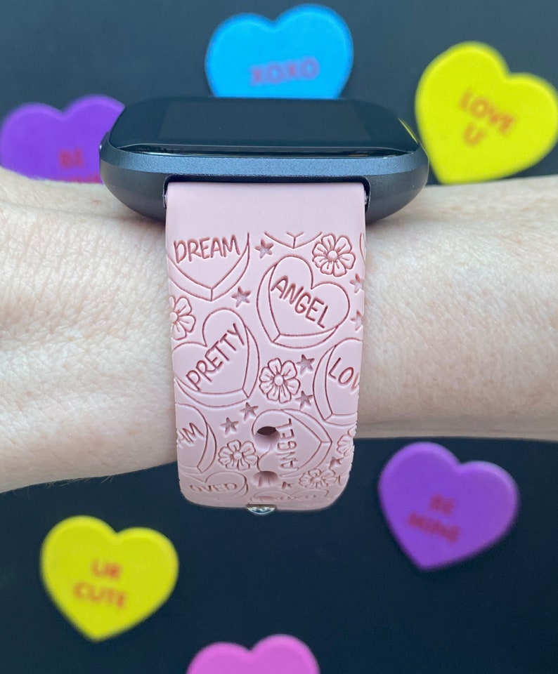 May include: A pink smartwatch strap with a heart design, featuring the words "Dream," "Angel," and "Pretty." The strap has floral and star details. Around the watch are colourful heart-shaped sweets with phrases such as "XOXO" and "Be Mine."