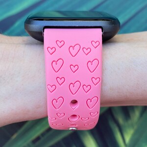 May include: A pink silicone watch strap with a heart pattern. The strap is designed for a smartwatch and has multiple holes for adjusting the fit.