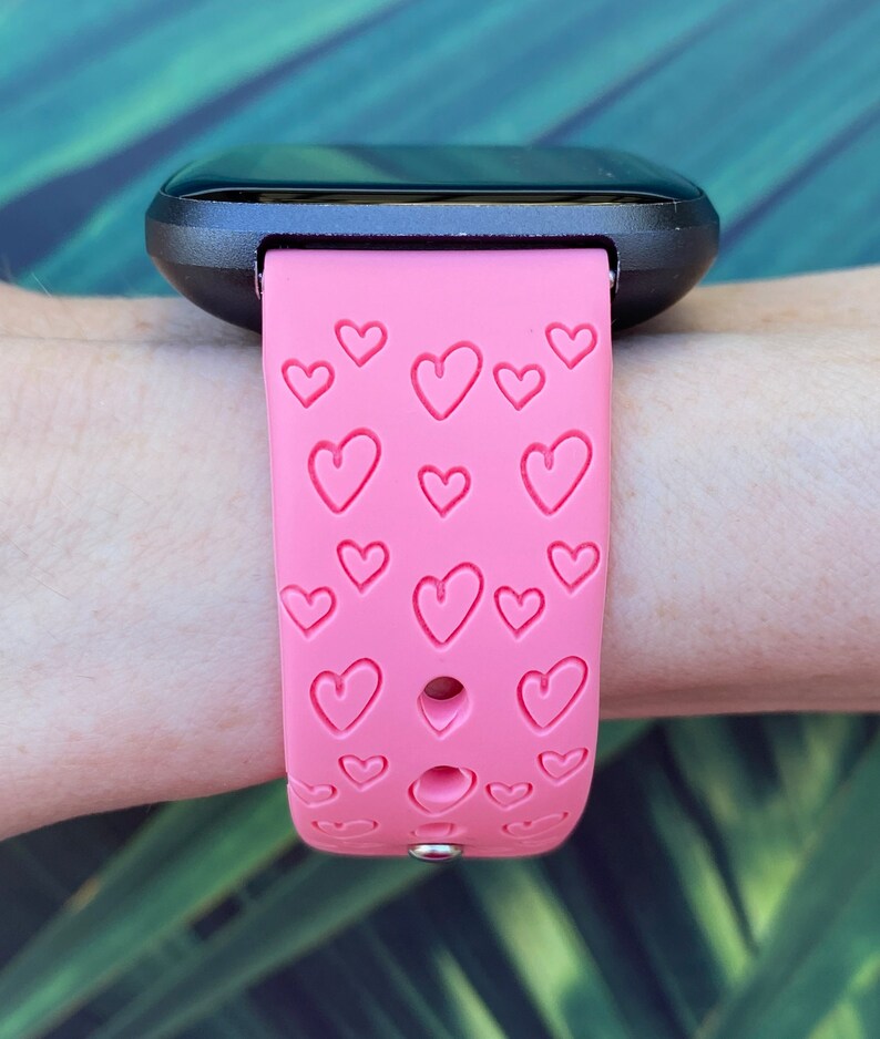May include: A pink silicone watch strap with a heart pattern. The strap is designed for a smartwatch and has multiple holes for adjusting the fit.