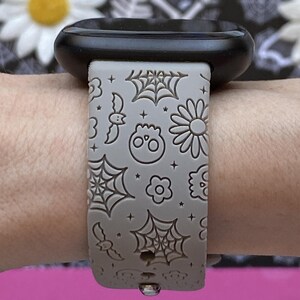 May include: A gray watch band with a Halloween theme. The band features a repeating pattern of spiderwebs, bats, skulls, and flowers. The watch face is black.
