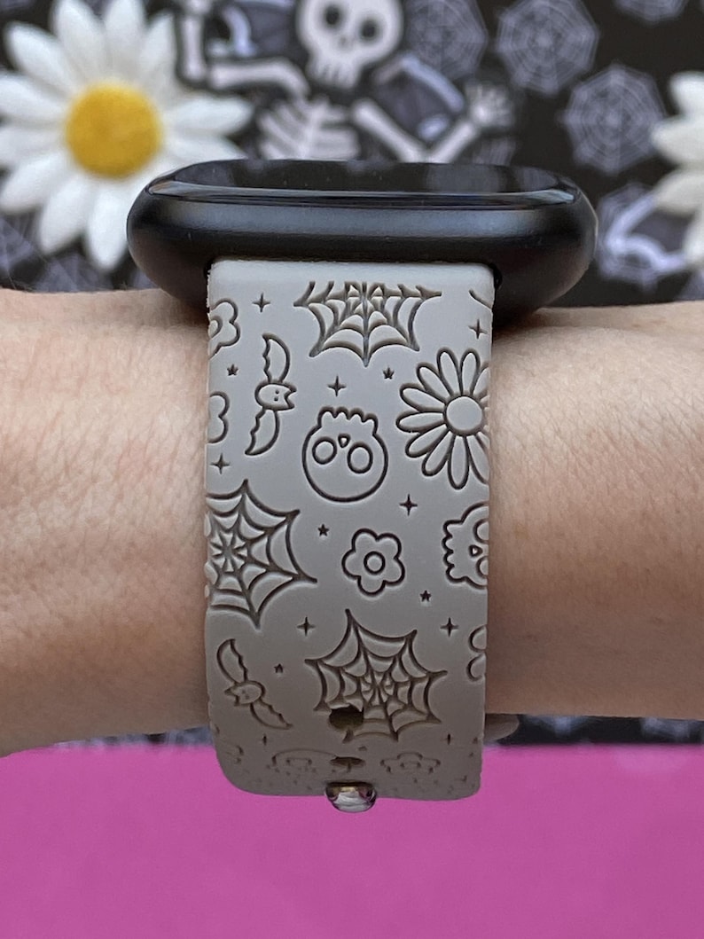 May include: A gray watch band with a Halloween theme. The band features a repeating pattern of spiderwebs, bats, skulls, and flowers. The watch face is black.