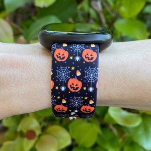 K&ouml;nnte beinhalten: Eine schwarze Smartwatch mit einem Halloween-Armband. Das Armband zeigt ein sich wiederholendes Muster aus orangefarbenen K&uuml;rbissen, Spinnweben und Maisbonbons auf schwarzem Hintergrund.