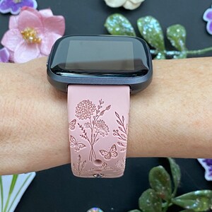 May include: A pink fitness tracker band with a floral design. The band features a delicate floral pattern with butterflies and leaves.
