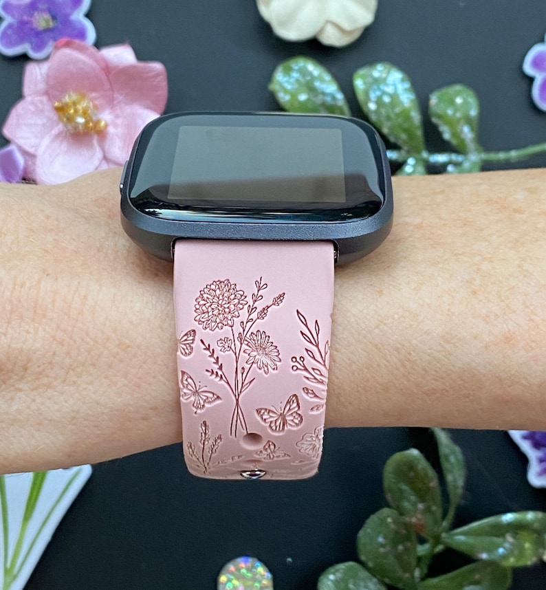May include: A pink fitness tracker band with a floral design. The band features a delicate floral pattern with butterflies and leaves.