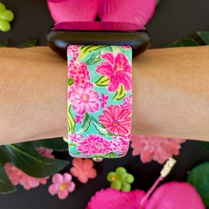 Op de afbeelding: Een zwarte smartwatch met een band met bloemenprint. De band heeft roze en groene bloemen op een lichtblauwe achtergrond.