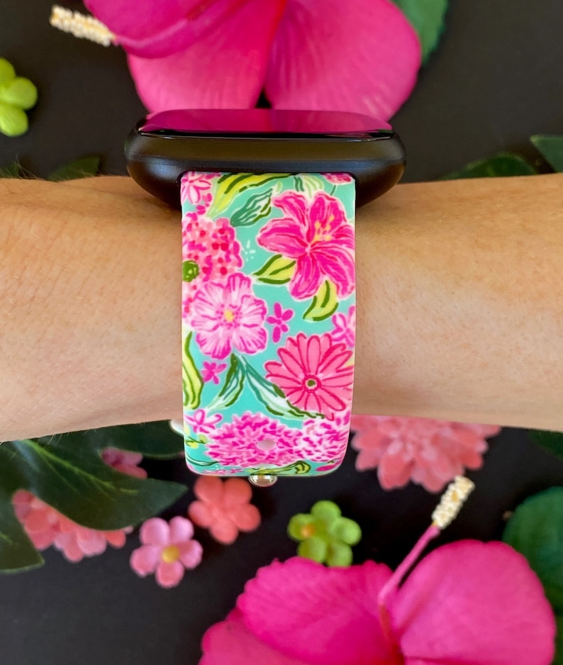 Op de afbeelding: Een zwarte smartwatch met een band met bloemenprint. De band heeft roze en groene bloemen op een lichtblauwe achtergrond.